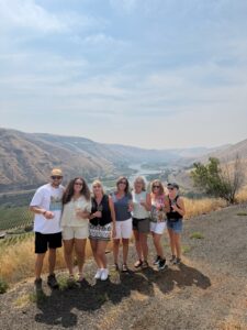 Seven adults pose with drinks on a hillside overlooking vineyards and a river valley in wine country, sunny day.