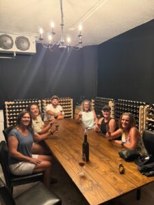 Six adults sit around a wooden table in a wine cellar, raising wine glasses with wine racks in the background.