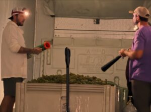 Two men stand on opposite sides of a chest filled with green cannabis buds, one holding a green and orange tool and the other a black bat-like object in a warehouse setting