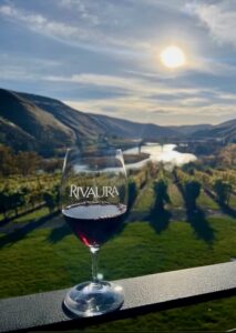 Wine glass with red wine on a balcony railing, etched with 'RIVAURA,' overlooking a vineyard and river valley at sunset