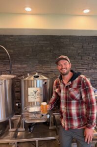 Smiling man in a red plaid shirt and cap holding a glass of beer next to stainless steel brewing tanks at River Ranch Co Brewing studio/facility.
