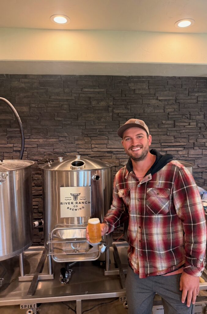 Smiling man in a red plaid shirt and cap holding a glass of beer next to stainless steel brewing tanks at River Ranch Co Brewing studio/facility.
