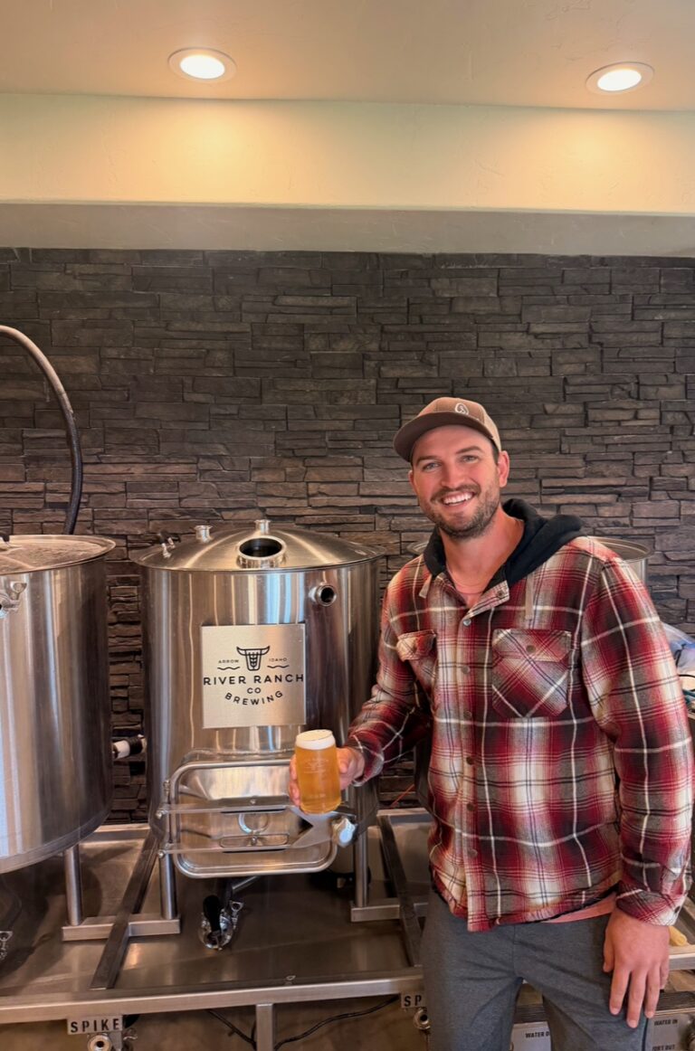 Smiling man in a red plaid shirt and cap holding a glass of beer next to stainless steel brewing tanks at River Ranch Co Brewing studio/facility.